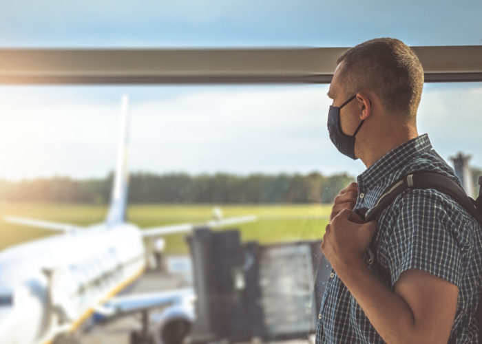 Man wearing a face mask and looking out an airport window at a plane on the tarmac
